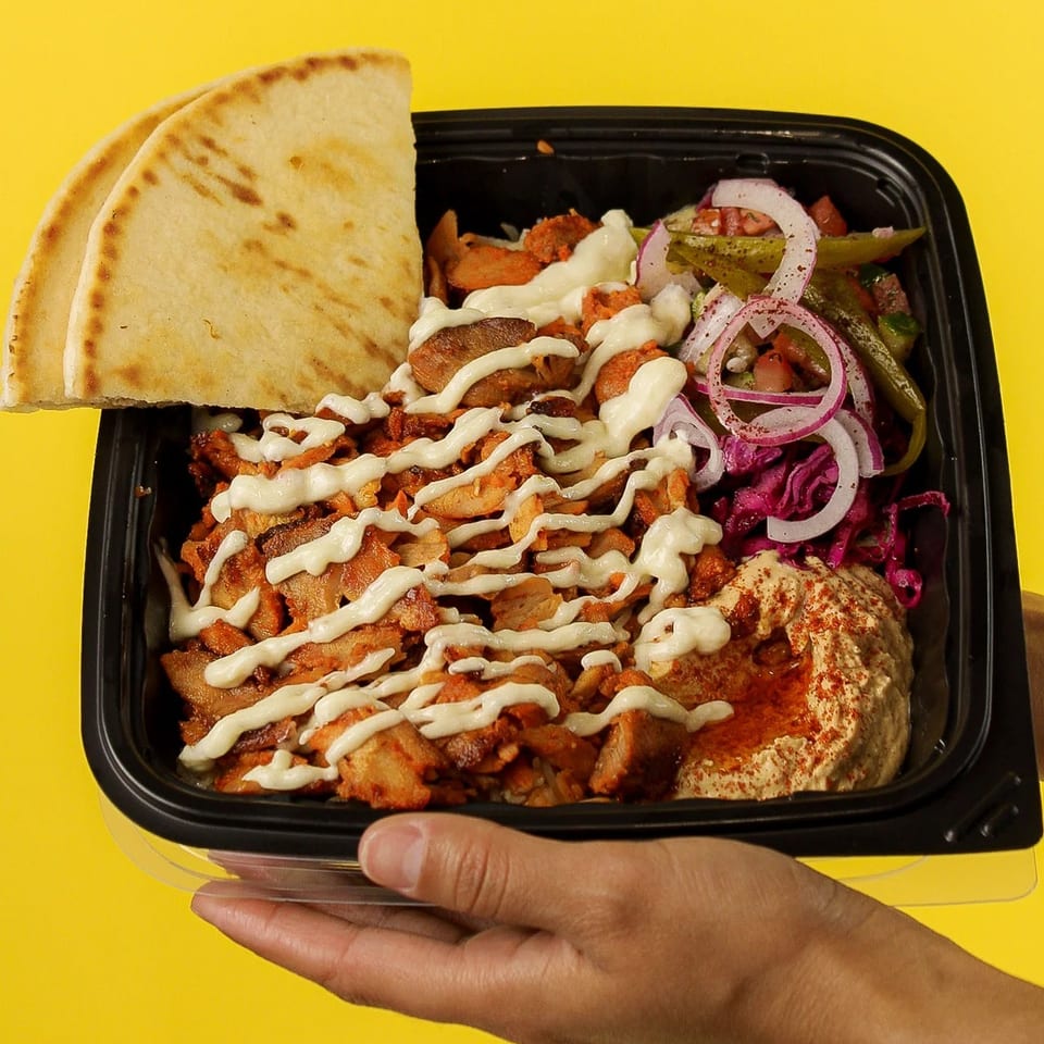 Takeout container with chicken shawarma, hummus, red cabbage, onions, pickles, and pita bread.