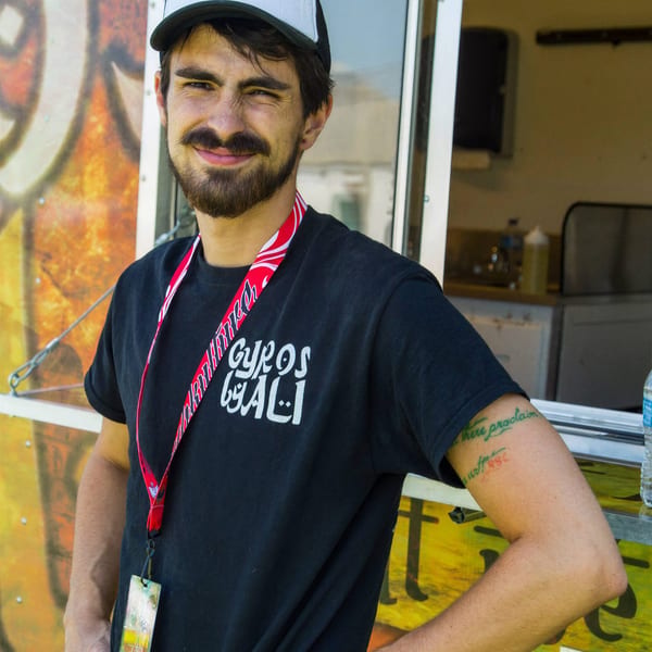 Smiling man in "Gyros by Ali" hat and t-shirt stands proudly in front of his food truck.