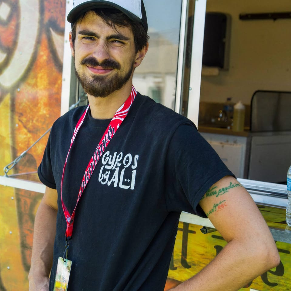 Smiling man in "Gyros by Ali" hat and t-shirt stands proudly in front of his food truck.
