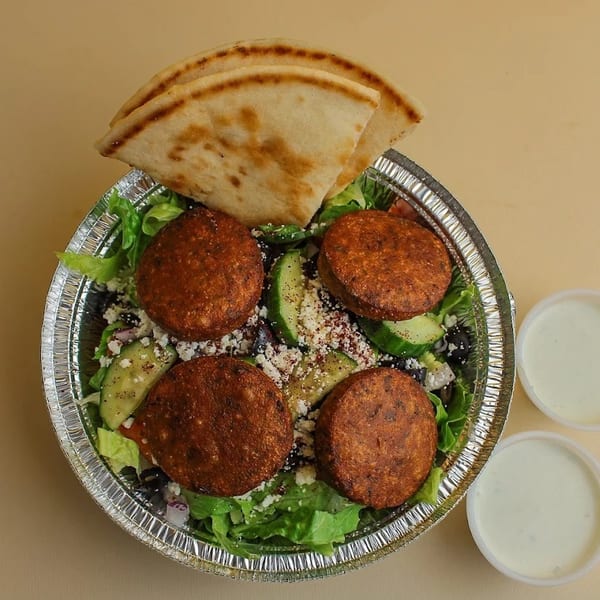 Falafel salad with fresh greens, cucumbers, feta, pita bread, and two sauce cups.