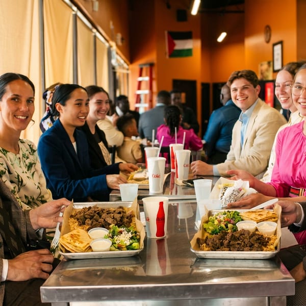Diverse group of people smiling and enjoying meals at a long table in a restaurant.