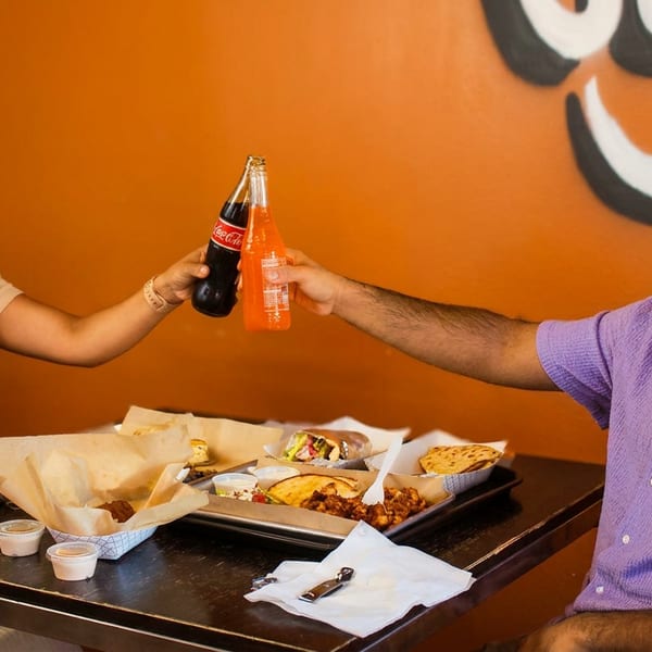 Two people clinking soda bottles over a table laden with various dishes in a restaurant.