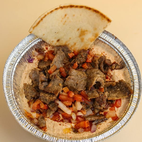 Foil container of hummus topped with sliced gyro meat, diced tomatoes, onions, and pita bread.