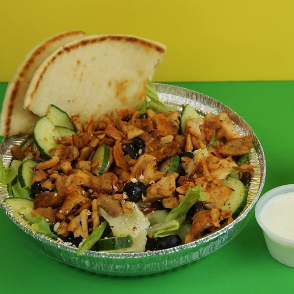 Chicken salad with pita bread, cucumbers, olives, and two sauce cups on a yellow and green background.