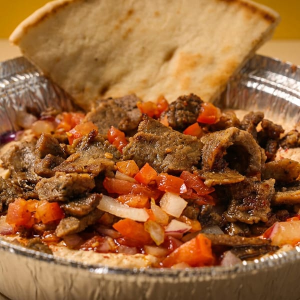 Hummus topped with gyro meat, diced tomatoes, and onions, served with pita bread in a foil container.