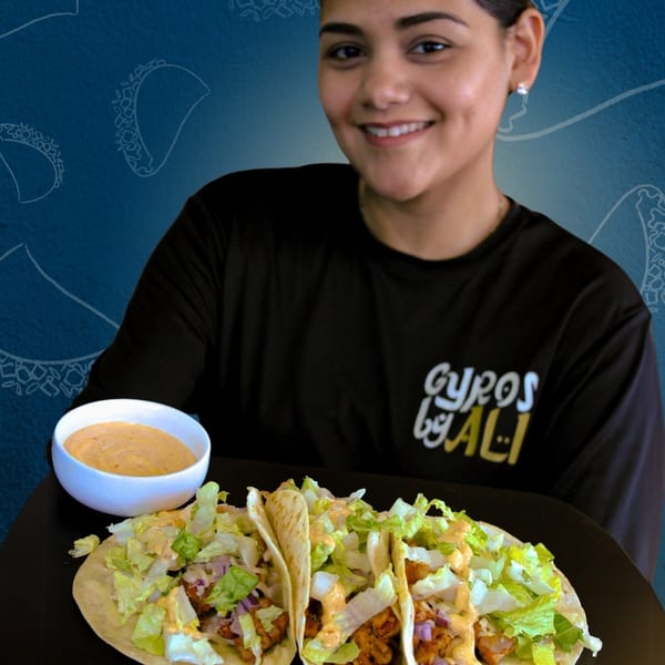 Smiling woman in "Gyros by Ali" shirt holding a tray with three tacos and a side of sauce.