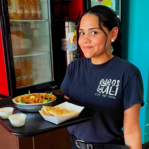 Smiling woman in "Gyros by Ali" shirt holds a tray with a gyro bowl, pita, and sauces.